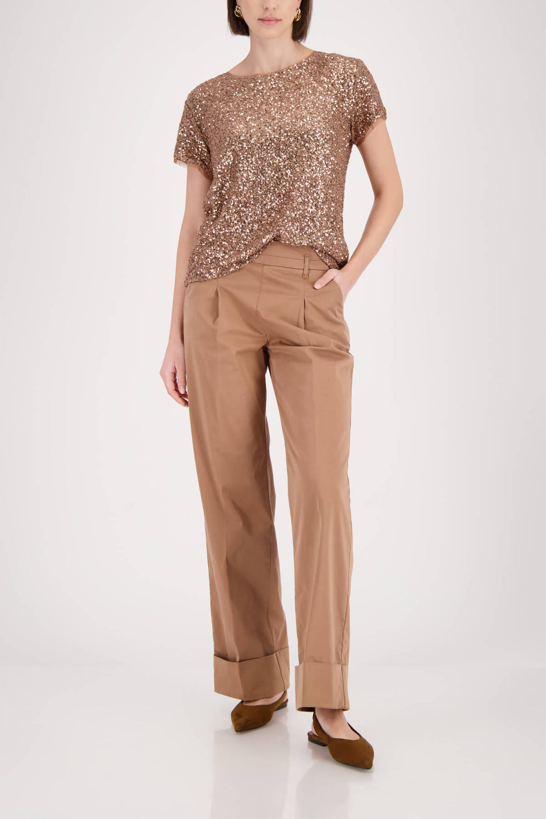 Monari modeling a short-sleeve crewneck top covered in shimmering rose gold sequins, paired with brown trousers, against a plain light gray studio background.
