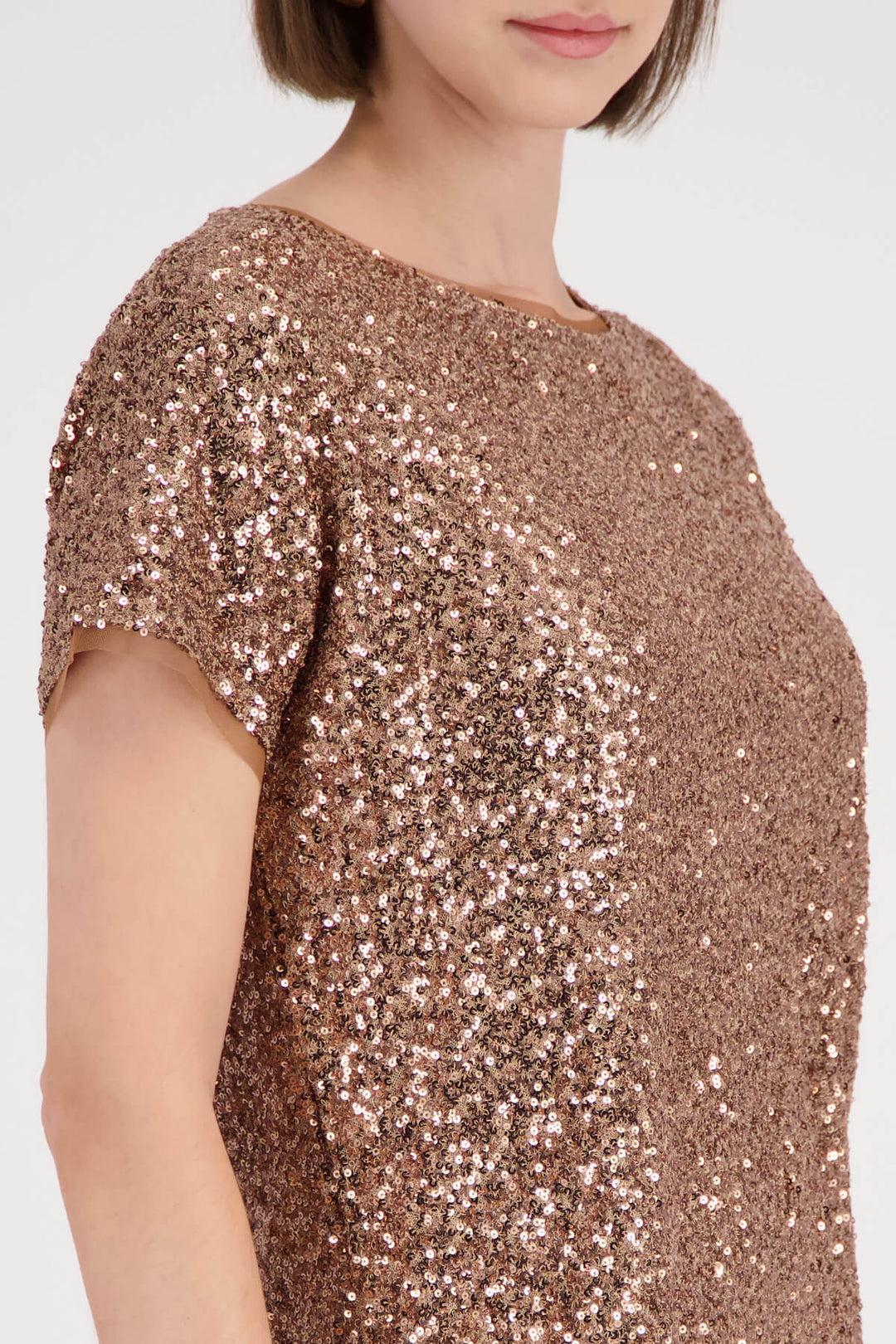 Monari modeling a short-sleeve crewneck top covered in shimmering rose gold sequins, paired with brown trousers, against a plain light gray studio background.