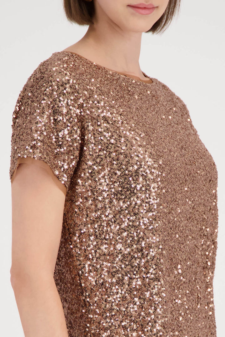 Monari modeling a short-sleeve crewneck top covered in shimmering rose gold sequins, paired with brown trousers, against a plain light gray studio background.
