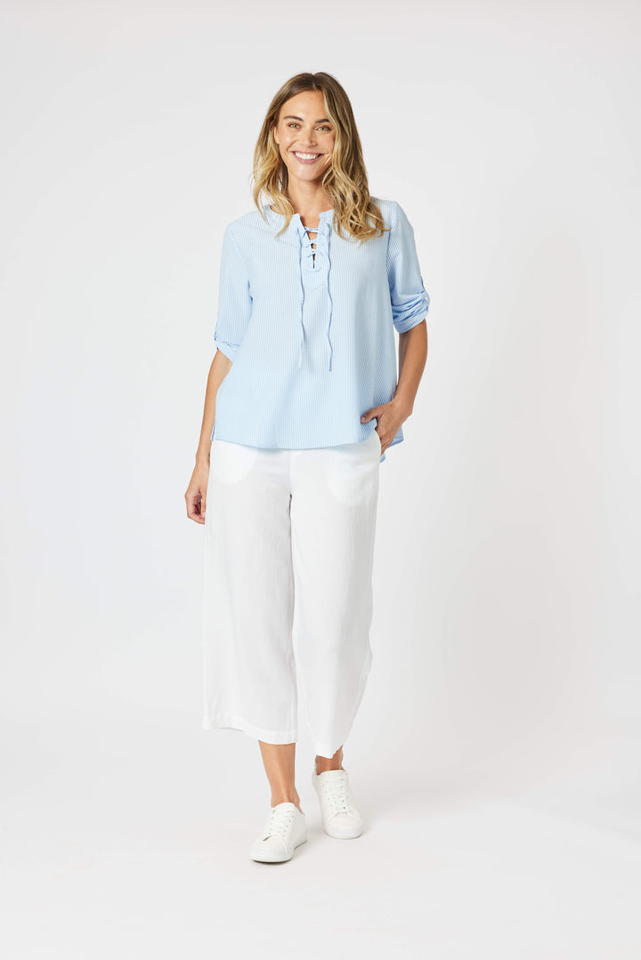 Gordon Smith Model A woman modeling a light blue long-sleeve blouse with a lace-up neckline and white cropped pants, standing against a plain white studio background.