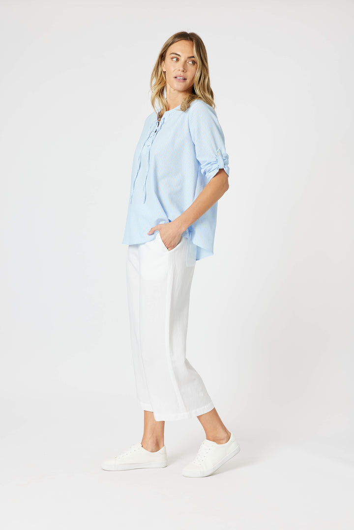 Gordon Smith Model A woman modeling a light blue long-sleeve blouse with a lace-up neckline and white cropped pants, standing against a plain white studio background.