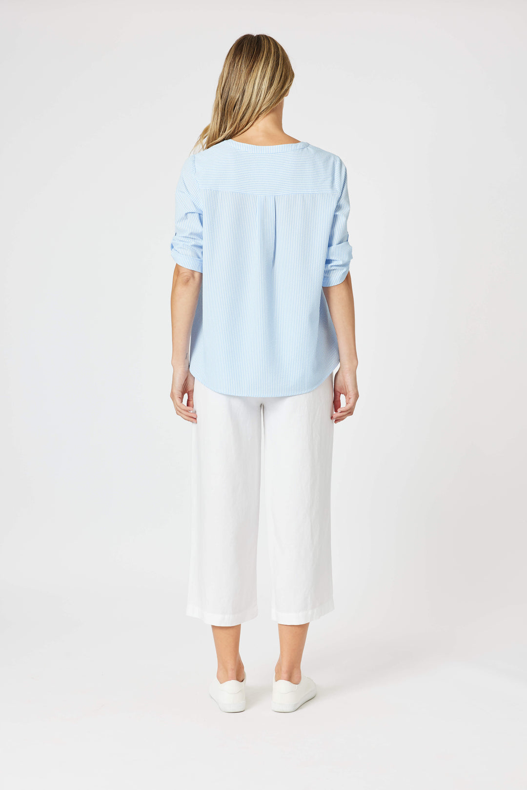 Gordon Smith Model A woman modeling a light blue long-sleeve blouse with a lace-up neckline and white cropped pants, standing against a plain white studio background.