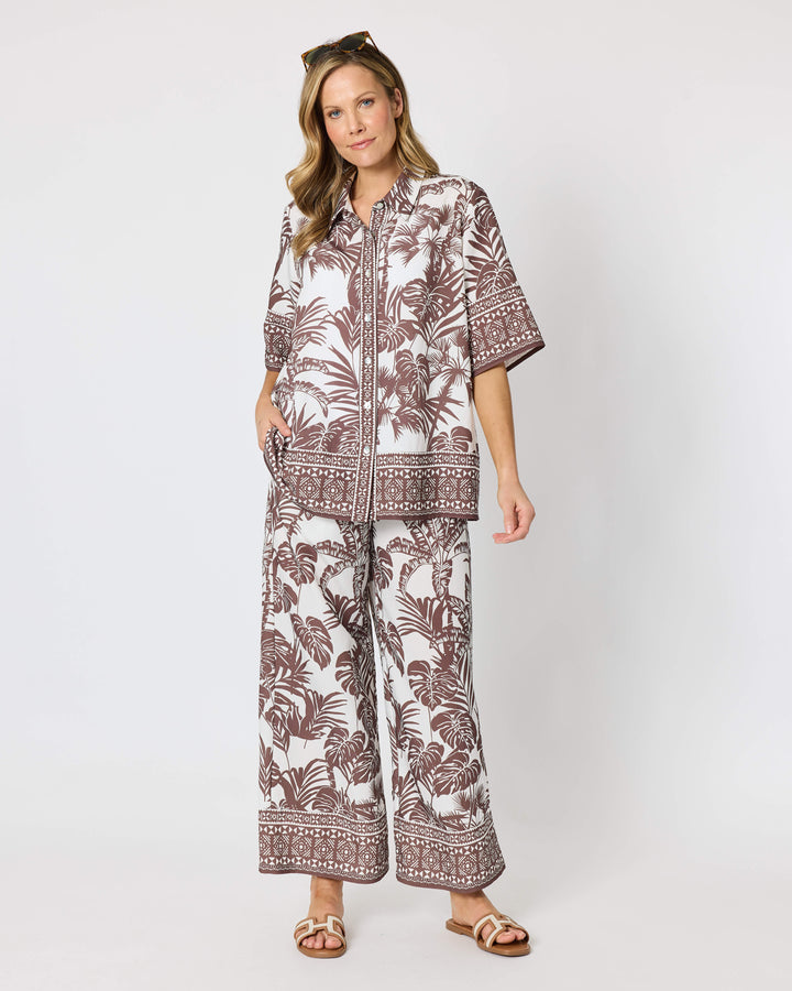 A model wearing a Hammock &amp; Vine matching two-piece lounge set featuring a brown palm leaf and geometric border print on an off-white background, paired with brown sandals against a plain light gray studio backdrop.