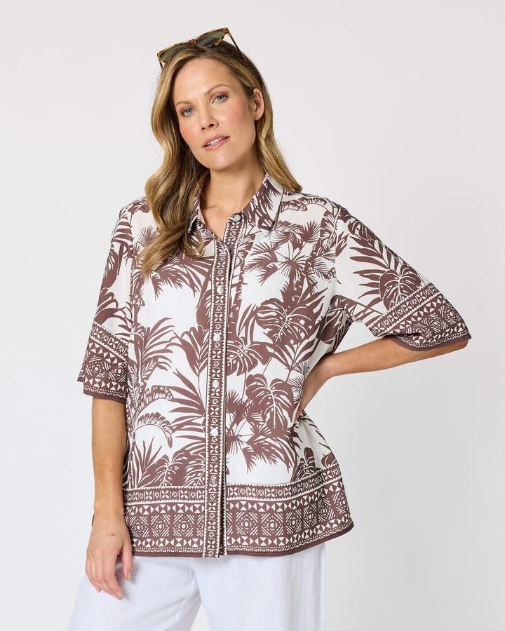 A hammock &amp; vine model wearing a short-sleeve button-up shirt with a brown and white tropical palm tree pattern, paired with white cropped pants and white slide sandals, against a plain white studio background.