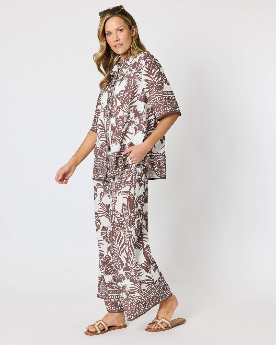 A model wearing a Hammock &amp; Vine matching two-piece lounge set featuring a brown palm leaf and geometric border print on an off-white background, paired with brown sandals against a plain light gray studio backdrop.
