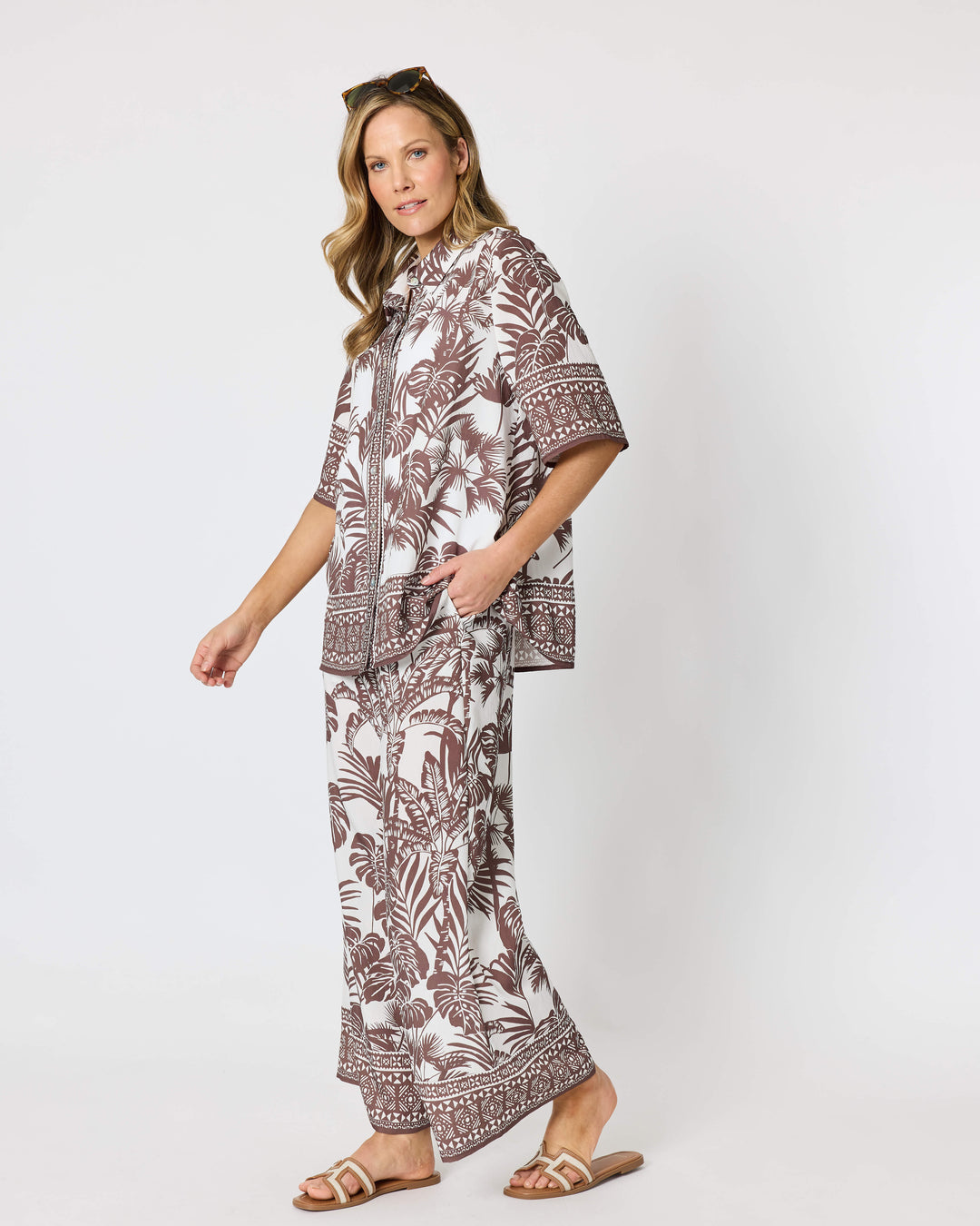 Hammock and Vine, modeling a matching two-piece lounge set featuring a brown and white tropical palm leaf pattern, consisting of a short-sleeve button-up shirt and wide-leg pants, styled with brown slide sandals against a plain white studio background.