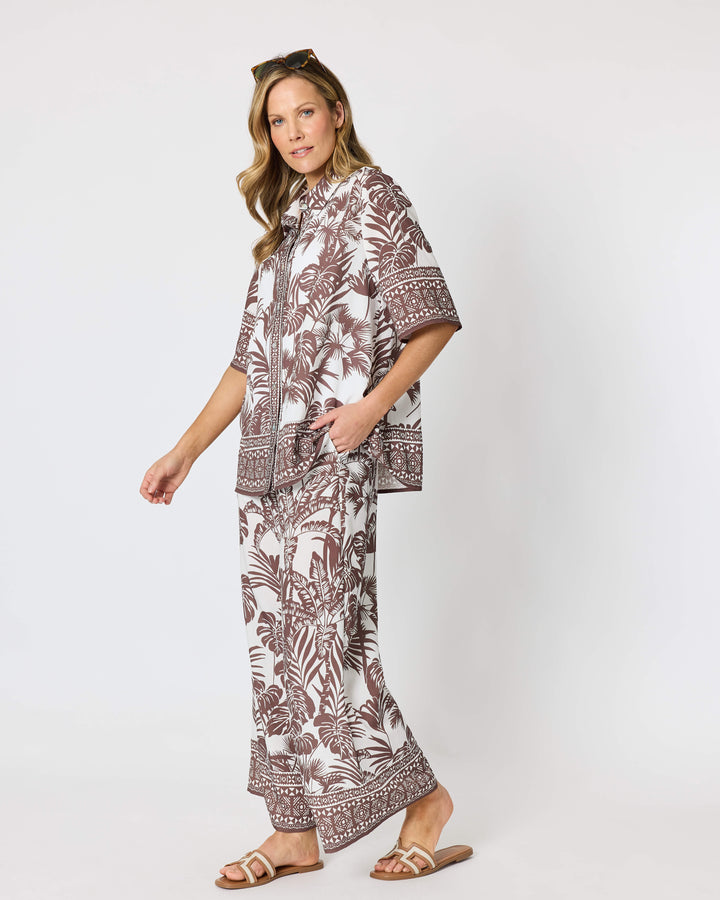 Hammock and Vine, modeling a matching two-piece lounge set featuring a brown and white tropical palm leaf pattern, consisting of a short-sleeve button-up shirt and wide-leg pants, styled with brown slide sandals against a plain white studio background.