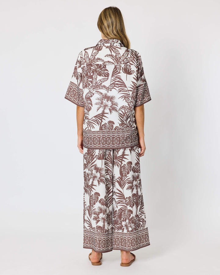 A model viewed from behind wearing a Hammock &amp; Vine matching two-piece lounge set with a brown tropical leaf and geometric border print on a white background, standing against a light gray studio backdrop.