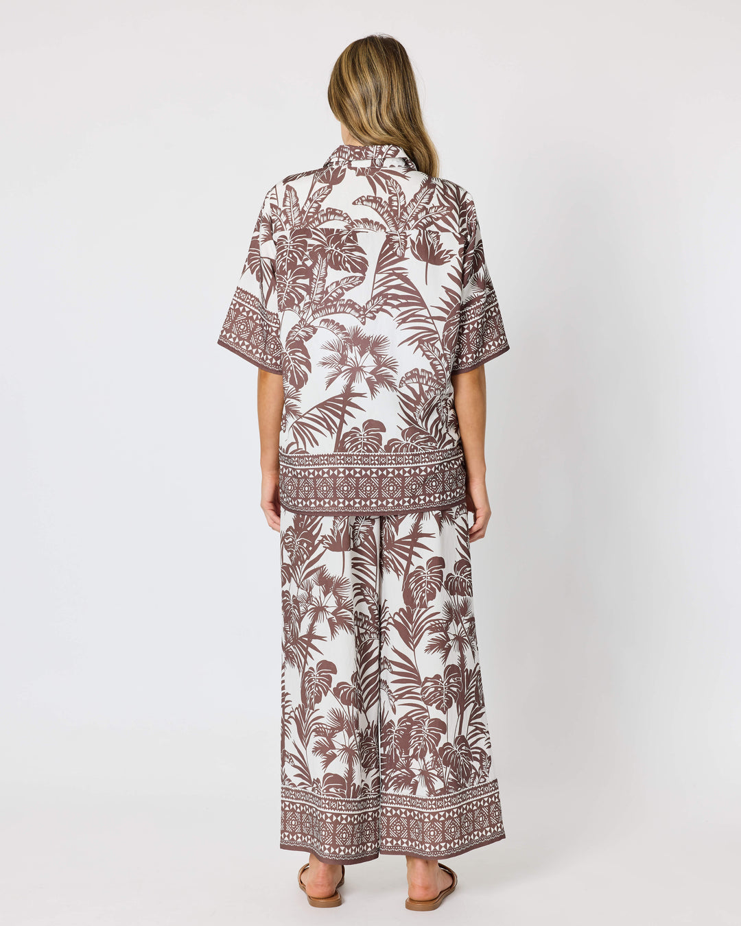 A model viewed from behind wearing a matching two-piece lounge set with a brown tropical leaf and geometric border print on a white background from Hammock and Vine, standing against a light gray studio backdrop.