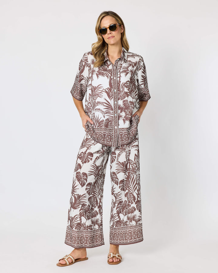 A model wearing a Hammock &amp; Vine matching two-piece lounge set featuring a brown palm leaf and geometric border print on an off-white background, paired with brown sandals against a plain light gray studio backdrop.