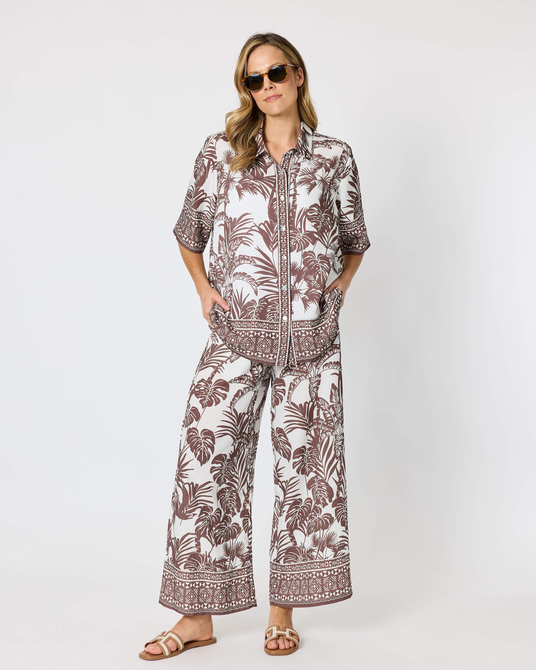 Hammock and Vine, modeling a matching two-piece lounge set featuring a brown and white tropical palm leaf pattern, consisting of a short-sleeve button-up shirt and wide-leg pants, styled with brown slide sandals against a plain white studio background.