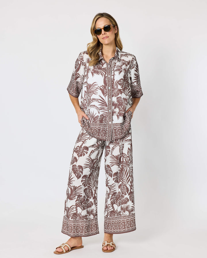 Hammock and Vine, modeling a matching two-piece lounge set featuring a brown and white tropical palm leaf pattern, consisting of a short-sleeve button-up shirt and wide-leg pants, styled with brown slide sandals against a plain white studio background.