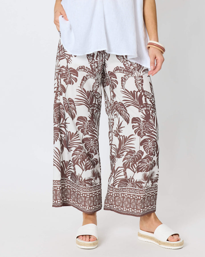 Hammock and Vine, modeling a white short-sleeve V-neck top with wide-leg brown and white tropical patterned pants, paired with white slide sandals, against a plain white studio background.