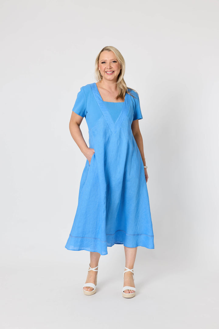 Gordon Smith woman  modeling a light blue midi dress with short sleeves, a square neckline, and side pockets, paired with white strappy sandals, against a plain white studio background.
