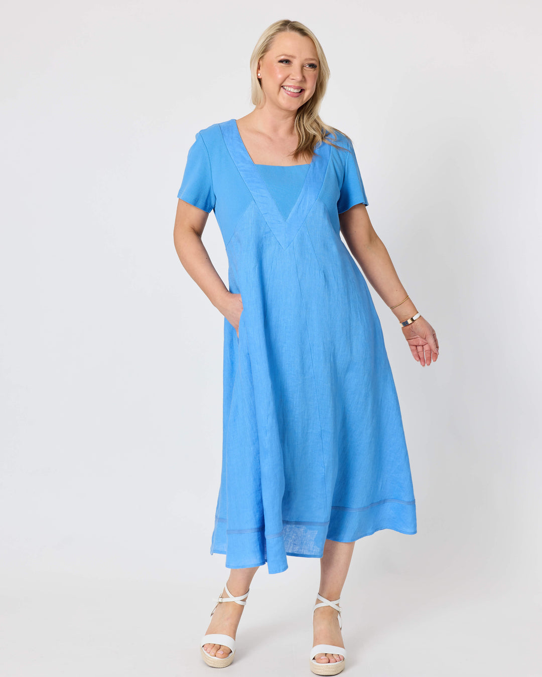 Gordon Smith woman  modeling a light blue midi dress with short sleeves, a square neckline, and side pockets, paired with white strappy sandals, against a plain white studio background.