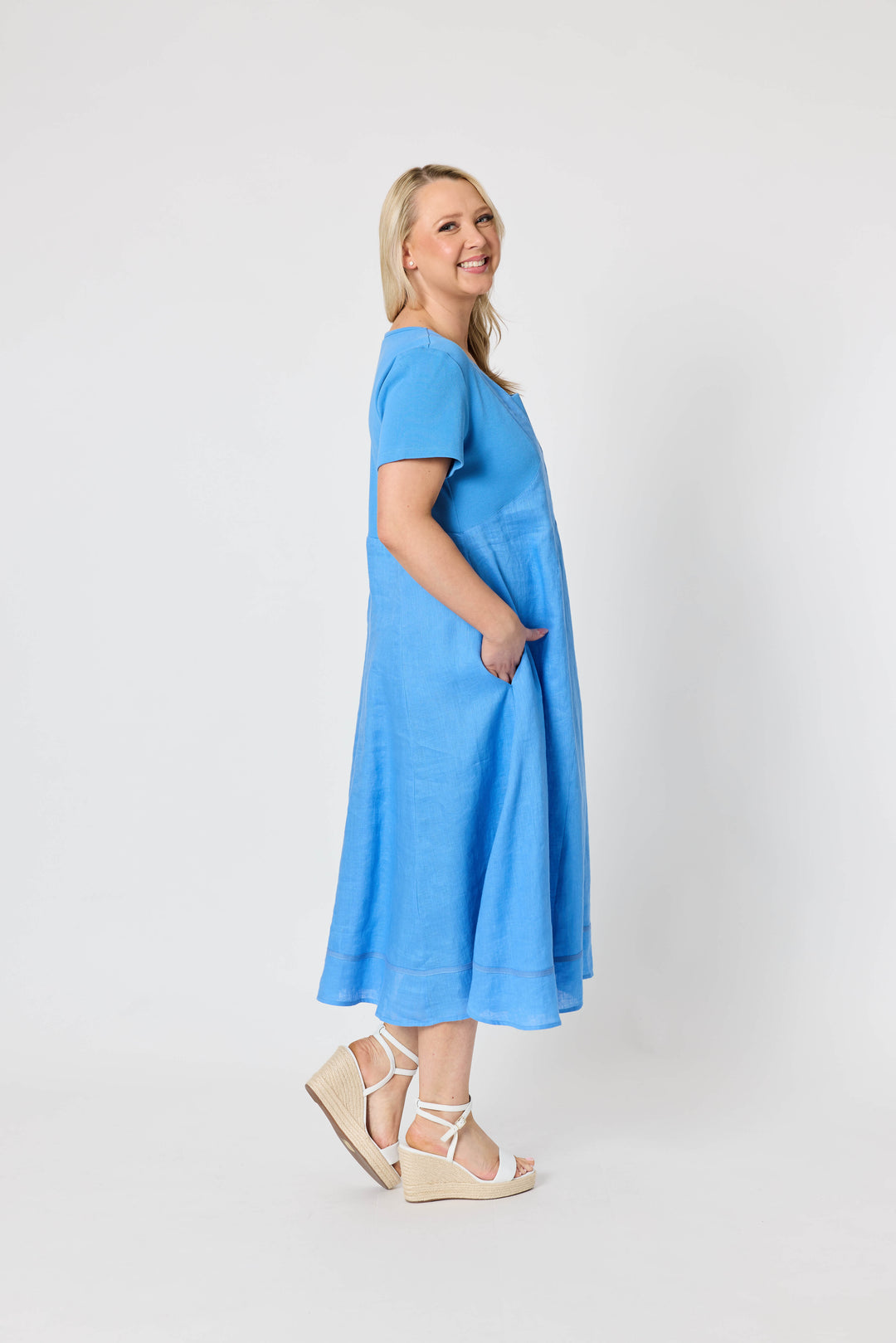 Gordon Smith woman  modeling a light blue midi dress with short sleeves, a square neckline, and side pockets, paired with white strappy sandals, against a plain white studio background.