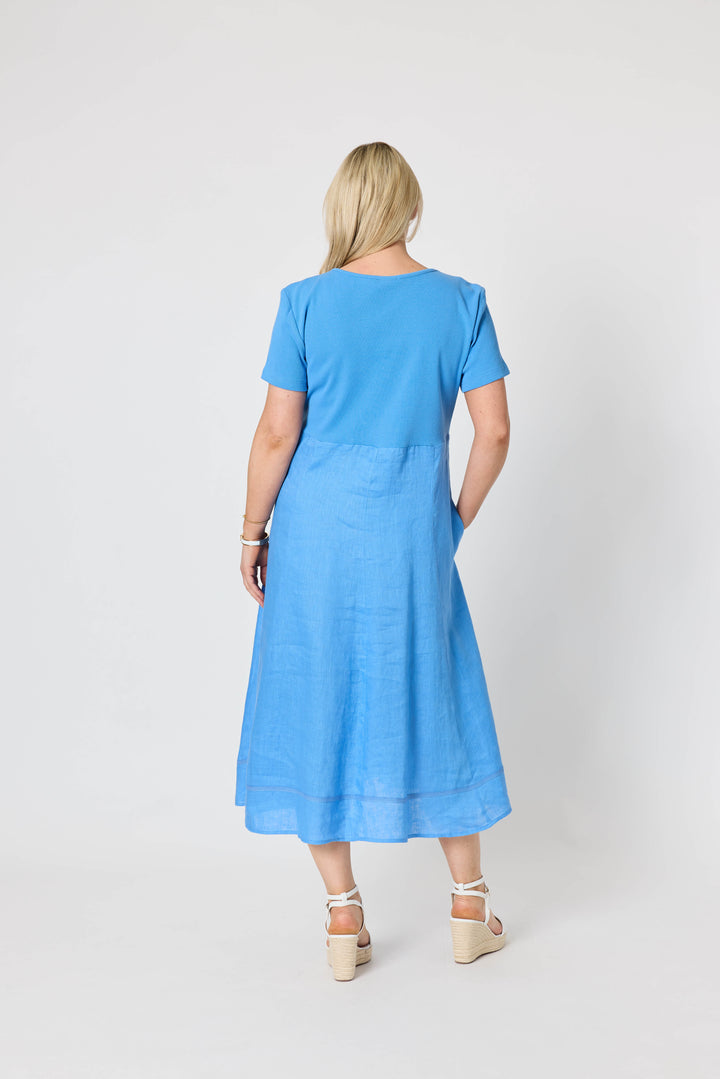 Gordon Smith woman  modeling a light blue midi dress with short sleeves, a square neckline, and side pockets, paired with white strappy sandals, against a plain white studio background. Back view