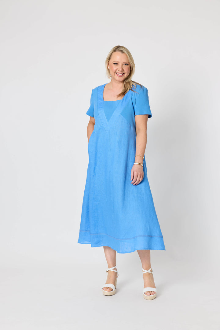 Gordon Smith woman  modeling a light blue midi dress with short sleeves, a square neckline, and side pockets, paired with white strappy sandals, against a plain white studio background.