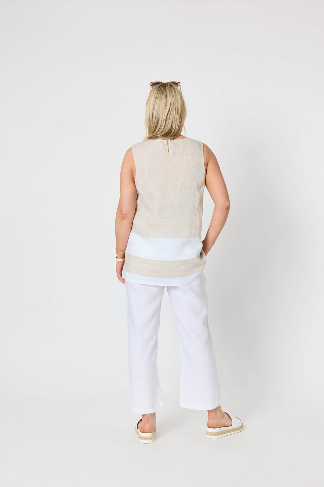 Gordon Smith woman modeling a sleeveless beige and white striped tunic top with white pants, standing against a plain light gray studio background. Back view