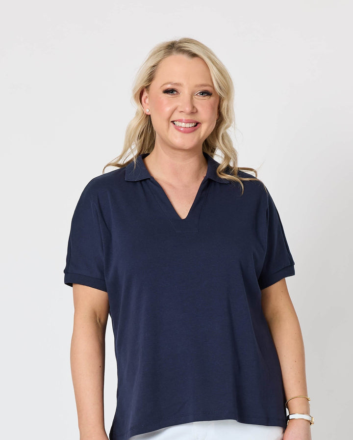 Gordon Smith Model A woman modeling a short-sleeve v-neck polo shirt in navy blue paired with white shorts and white sneakers, standing against a plain light gray studio background.