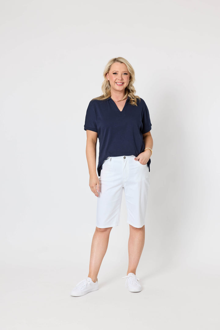 Gordon Smith Model A woman modeling a short-sleeve v-neck polo shirt in navy blue paired with white shorts and white sneakers, standing against a plain light gray studio background.
