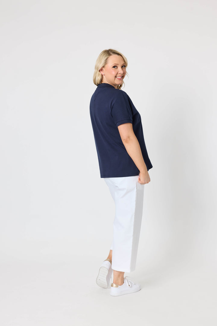 Gordon Smith Model A woman modeling a short-sleeve v-neck polo shirt in navy blue paired with white shorts and white sneakers, standing against a plain light gray studio background.