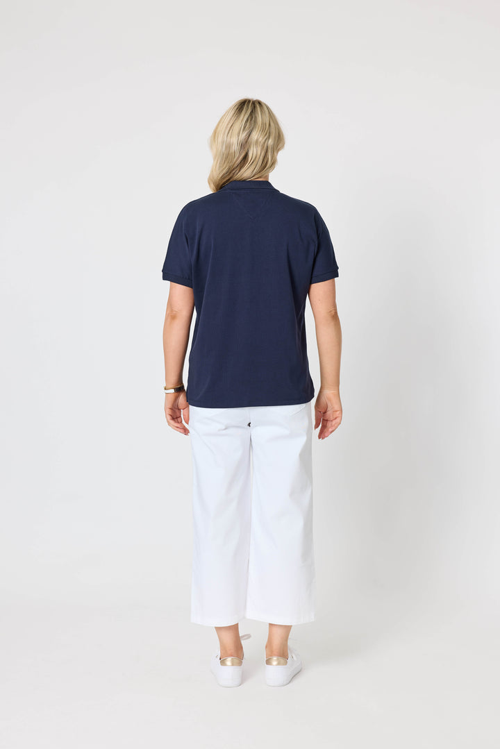 Gordon Smith Model A woman modeling a short-sleeve v-neck polo shirt in navy blue paired with white shorts and white sneakers, standing against a plain light gray studio background.