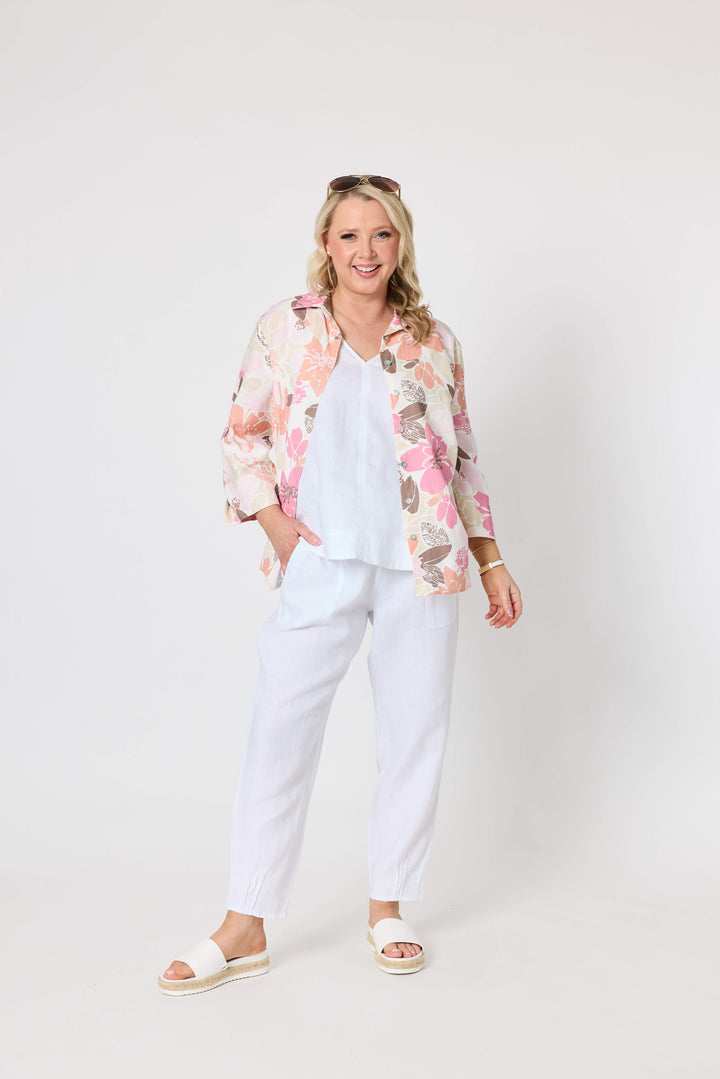 Gordon Smith woman modeling a long-sleeve button-up shirt with a pink, peach, and brown floral pattern on a cream background, paired with white pants and white slide sandals, against a plain light gray studio background.