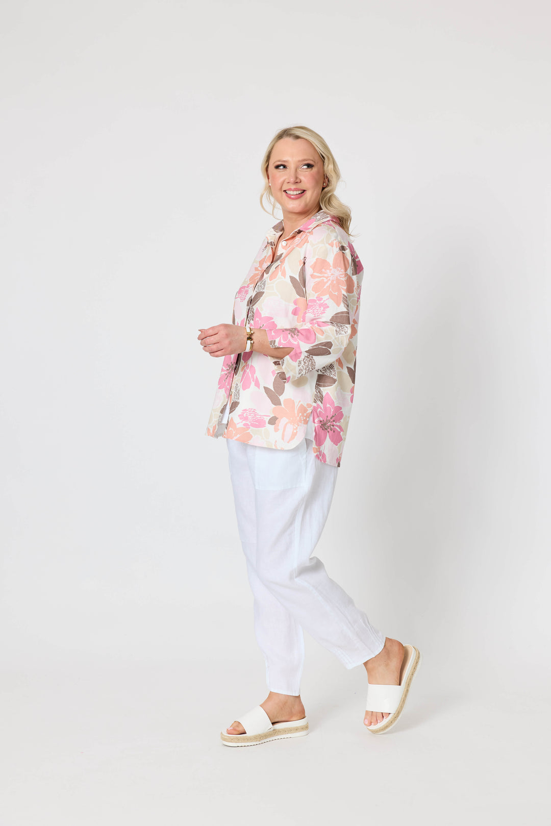 Gordon Smith woman modeling a long-sleeve button-up shirt with a pink, peach, and brown floral pattern on a cream background, paired with white pants and white slide sandals, against a plain light gray studio background.