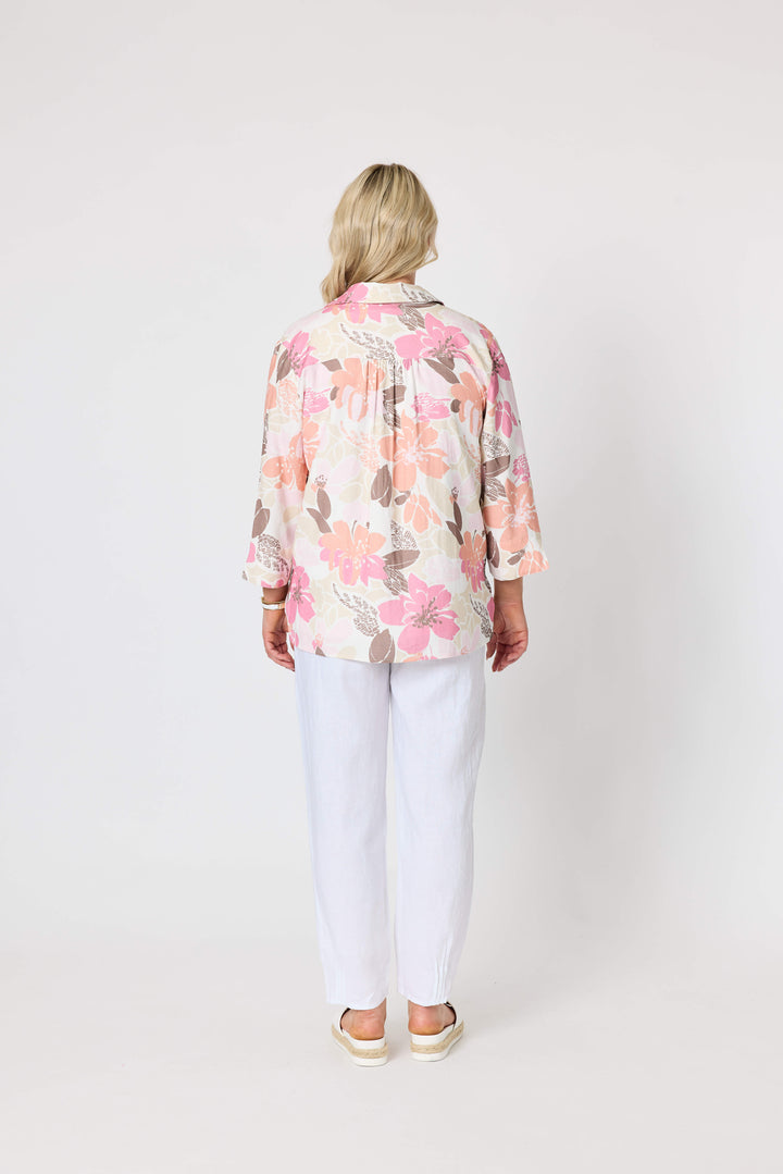 Gordon Smith woman modeling a long-sleeve button-up shirt with a pink, peach, and brown floral pattern on a cream background, paired with white pants and white slide sandals, against a plain light gray studio background. Back view