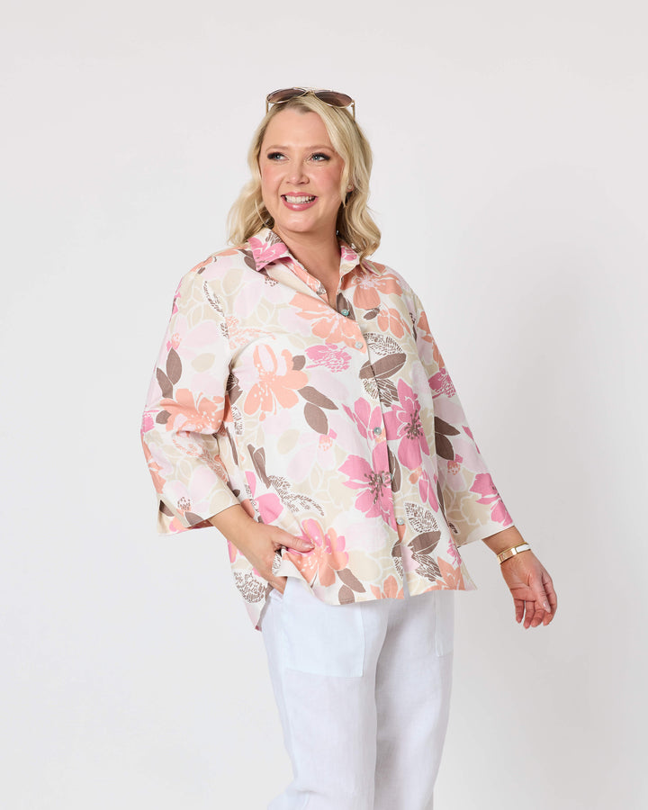 Gordon Smith woman modeling a long-sleeve button-up shirt with a pink, peach, and brown floral pattern on a cream background, paired with white pants and white slide sandals, against a plain light gray studio background.