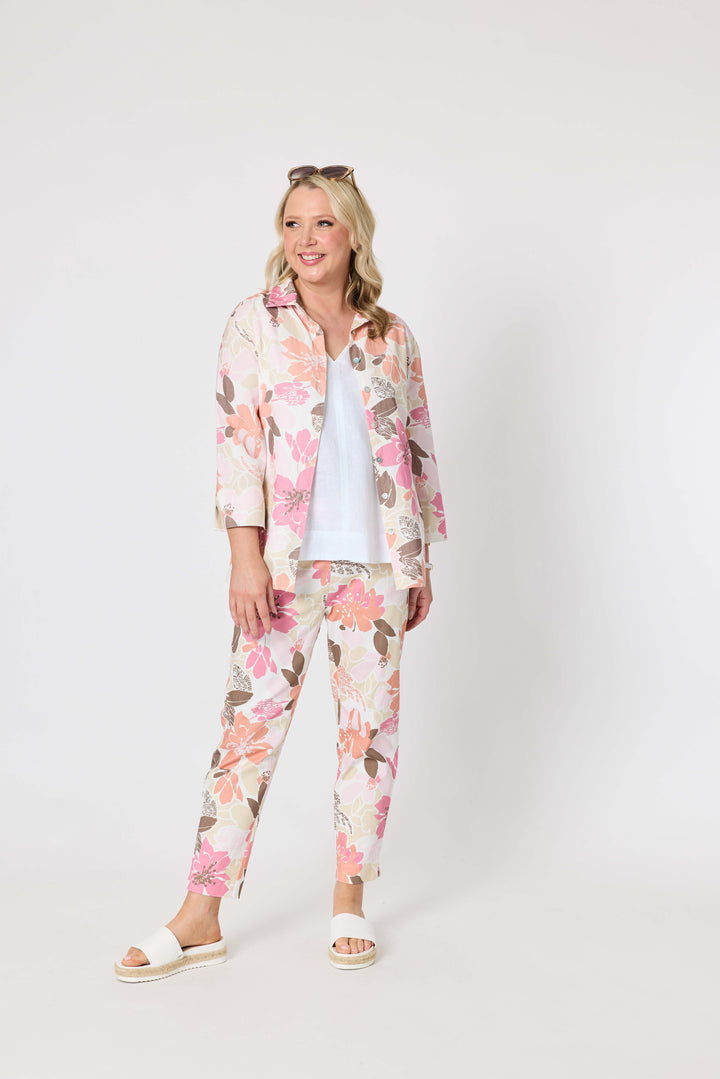 Gordon Smith woman modeling a long-sleeve button-up shirt with a pink, peach, and brown floral pattern on a cream background, paired with white pants and white slide sandals, against a plain light gray studio background.