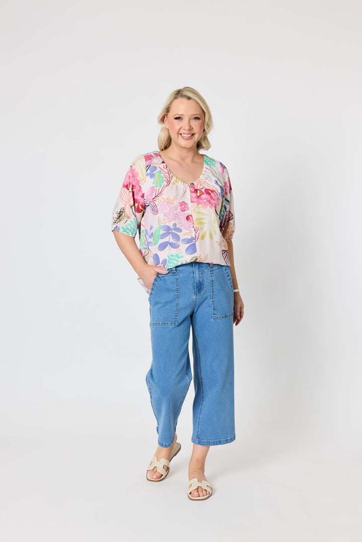 Gordon Smith Model A woman modeling a short-sleeve V-neck blouse with a colorful floral and botanical print, paired with blue cropped jeans and beige slide sandals, against a plain light gray studio background.