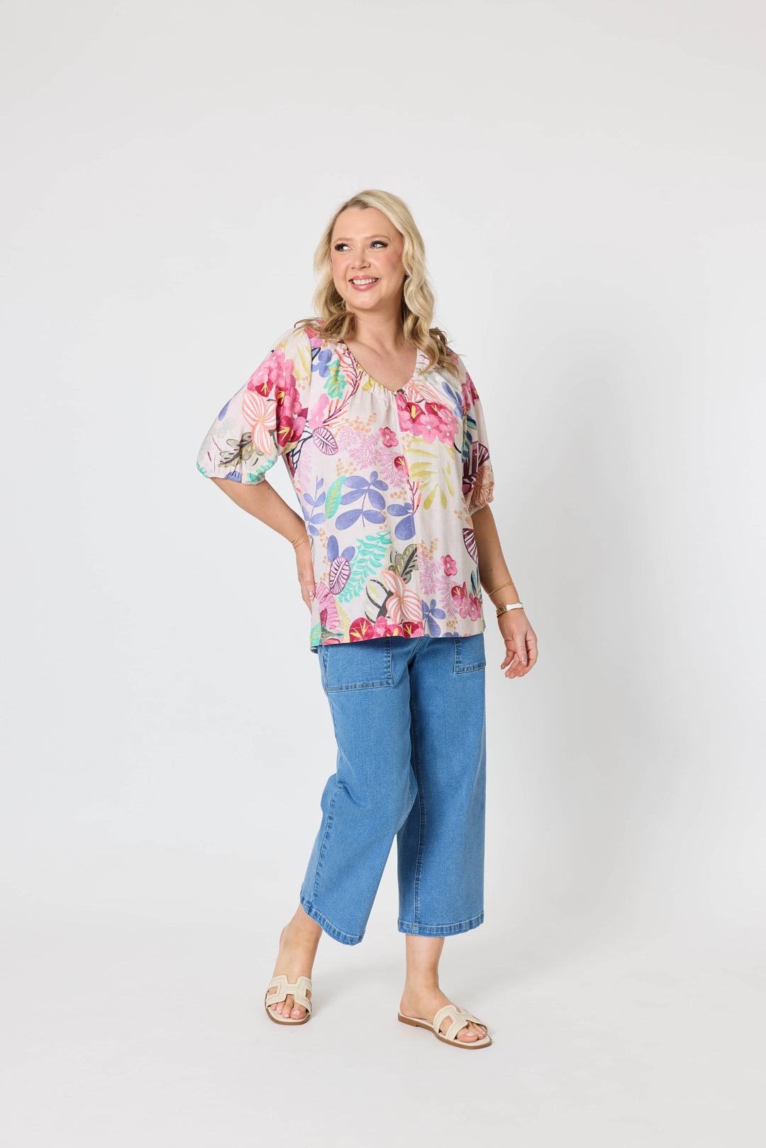 Gordon Smith Model A woman modeling a short-sleeve V-neck blouse with a colorful floral and botanical print, paired with blue cropped jeans and beige slide sandals, against a plain light gray studio background.