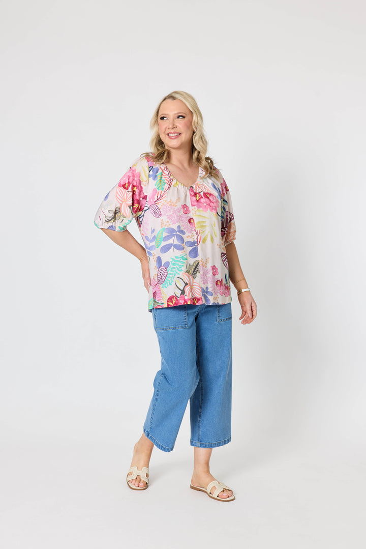 Gordon Smith Model A woman modeling a short-sleeve V-neck blouse with a colorful floral and botanical print, paired with blue cropped jeans and beige slide sandals, against a plain light gray studio background.