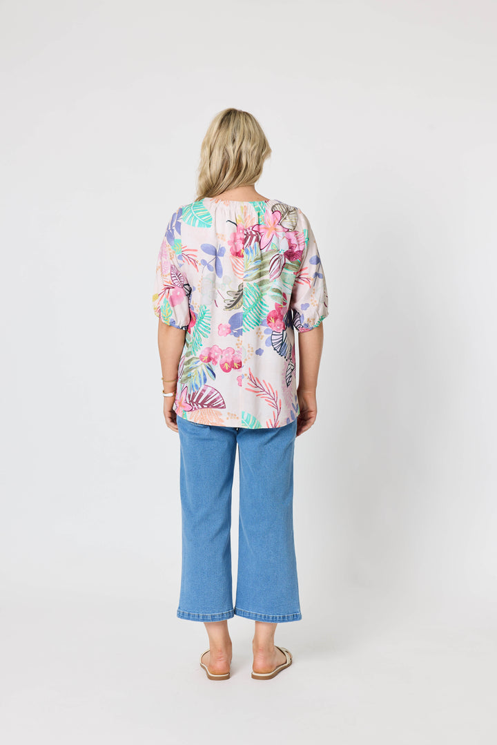 Gordon Smith Model A woman modeling a short-sleeve V-neck blouse with a colorful floral and botanical print, paired with blue cropped jeans and beige slide sandals, against a plain light gray studio background.