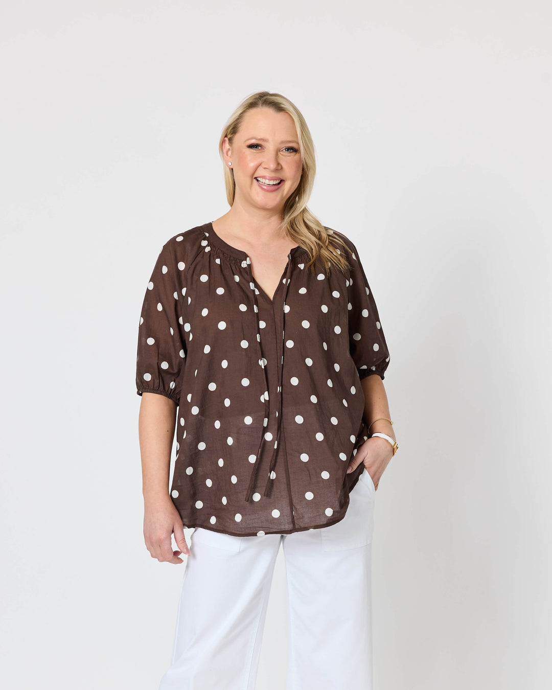 Gordon Smith modeling a brown short-sleeve button-up blouse with white polka dots, paired with white cropped pants and white slide sandals, against a plain light gray studio background.