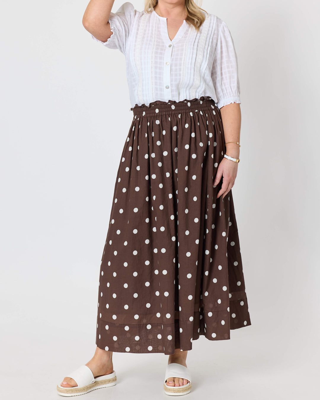 Gordon Smith modeling a white short-sleeve button-up shirt and a brown high-waisted maxi skirt with white polka dots, paired with white slide sandals, against a plain white studio background.