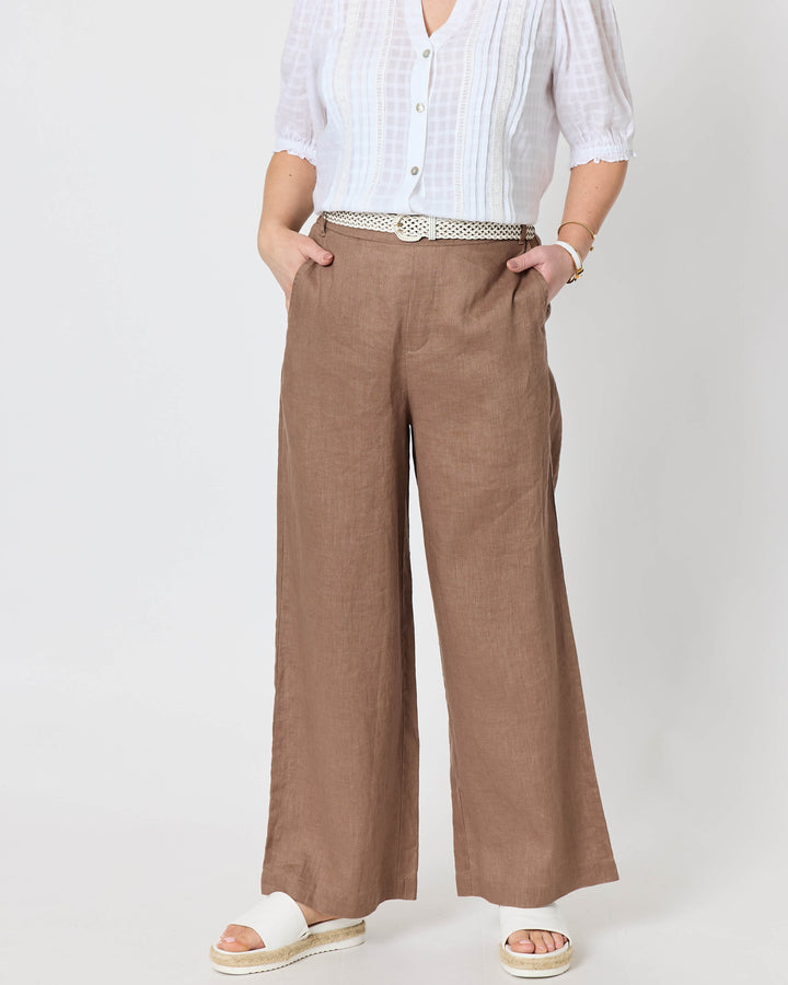 Gordon Smith modeling a white and blue striped short-sleeve button-up shirt with wide-leg brown trousers, a white belt, and white slide sandals, standing against a plain white studio background.