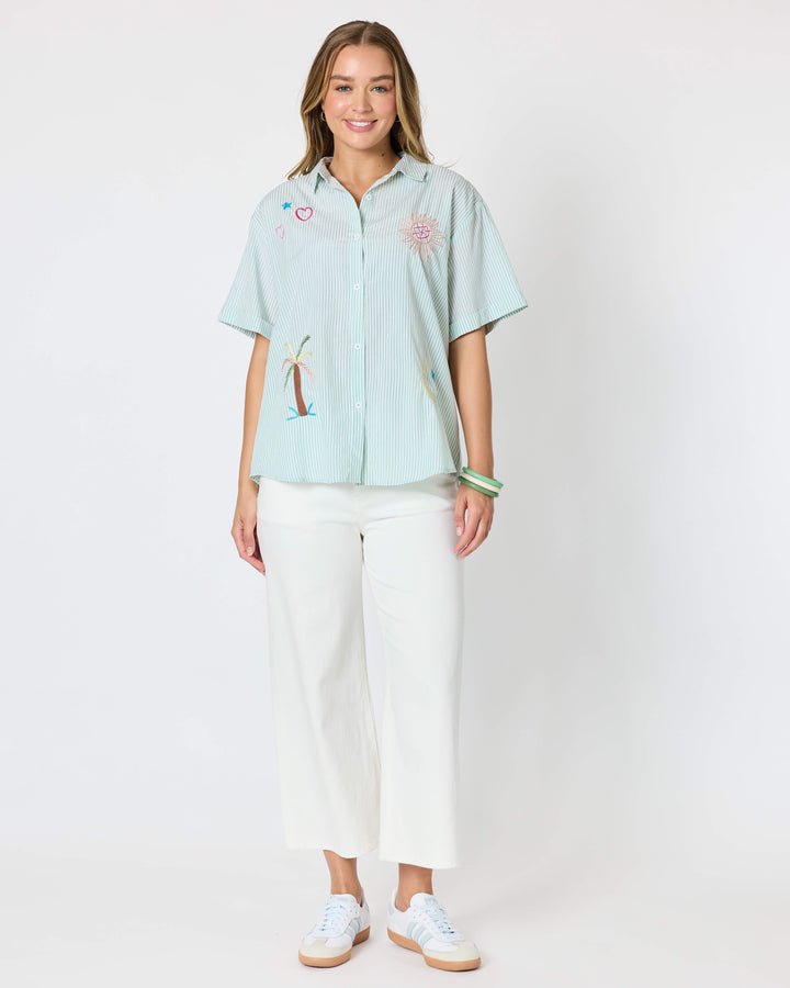 Threadz Model A woman modeling a light mint green short-sleeve button-up shirt with colorful embroidered details, paired with white cropped pants and white sneakers, against a plain white studio background.