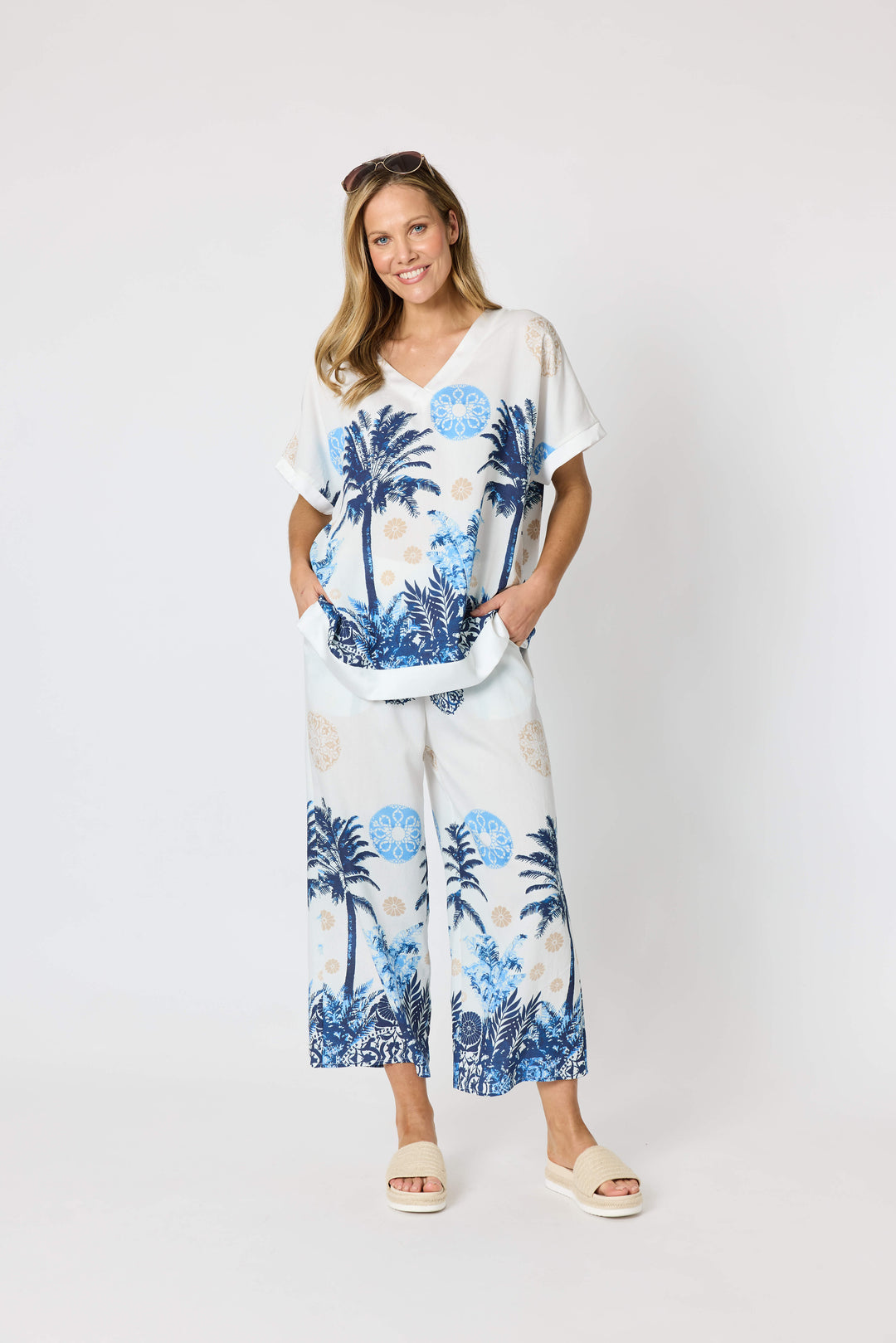 Clarity  woman modeling a matching two-piece lounge set featuring a white short-sleeve top and cropped pants with a blue tropical palm tree print, styled with beige slide sandals against a plain white studio background.