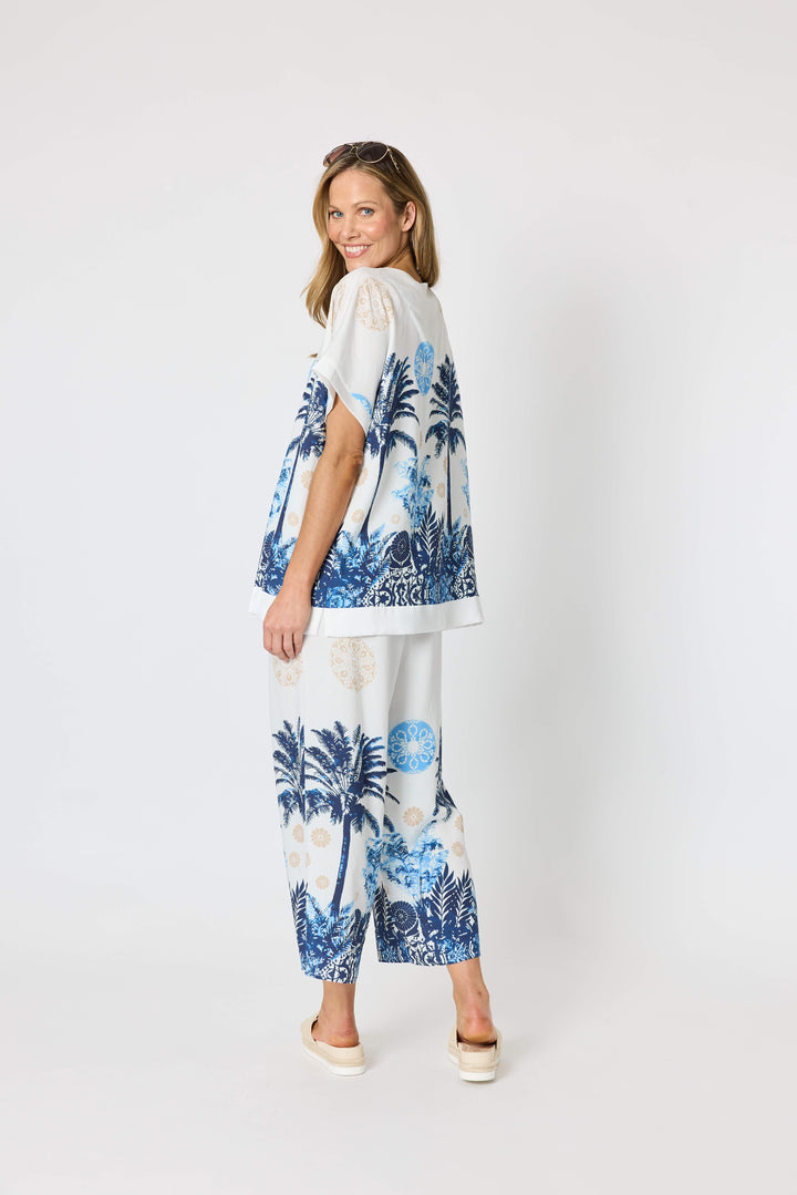 Clarity  woman modeling a two-piece lounge set consisting of a white short-sleeve top with a blue palm tree graphic and matching white wide-leg pants with a blue palm tree and beige sun motif, set against a plain white studio background.
Back view