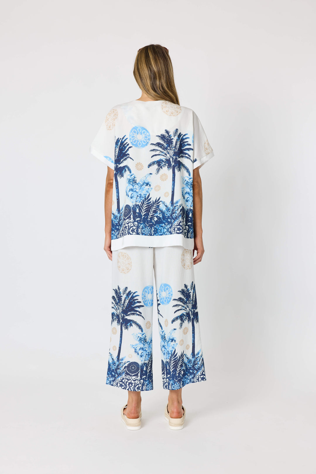 Clarity  woman modeling a matching two-piece lounge set featuring a white short-sleeve top and cropped pants with a blue tropical palm tree print, styled with beige slide sandals against a plain white studio background. Back view