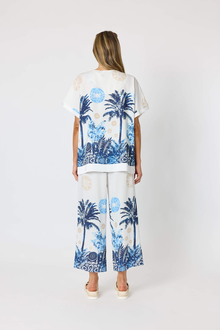 Clarity  woman modeling a matching two-piece lounge set featuring a white short-sleeve top and cropped pants with a blue tropical palm tree print, styled with beige slide sandals against a plain white studio background. Back view