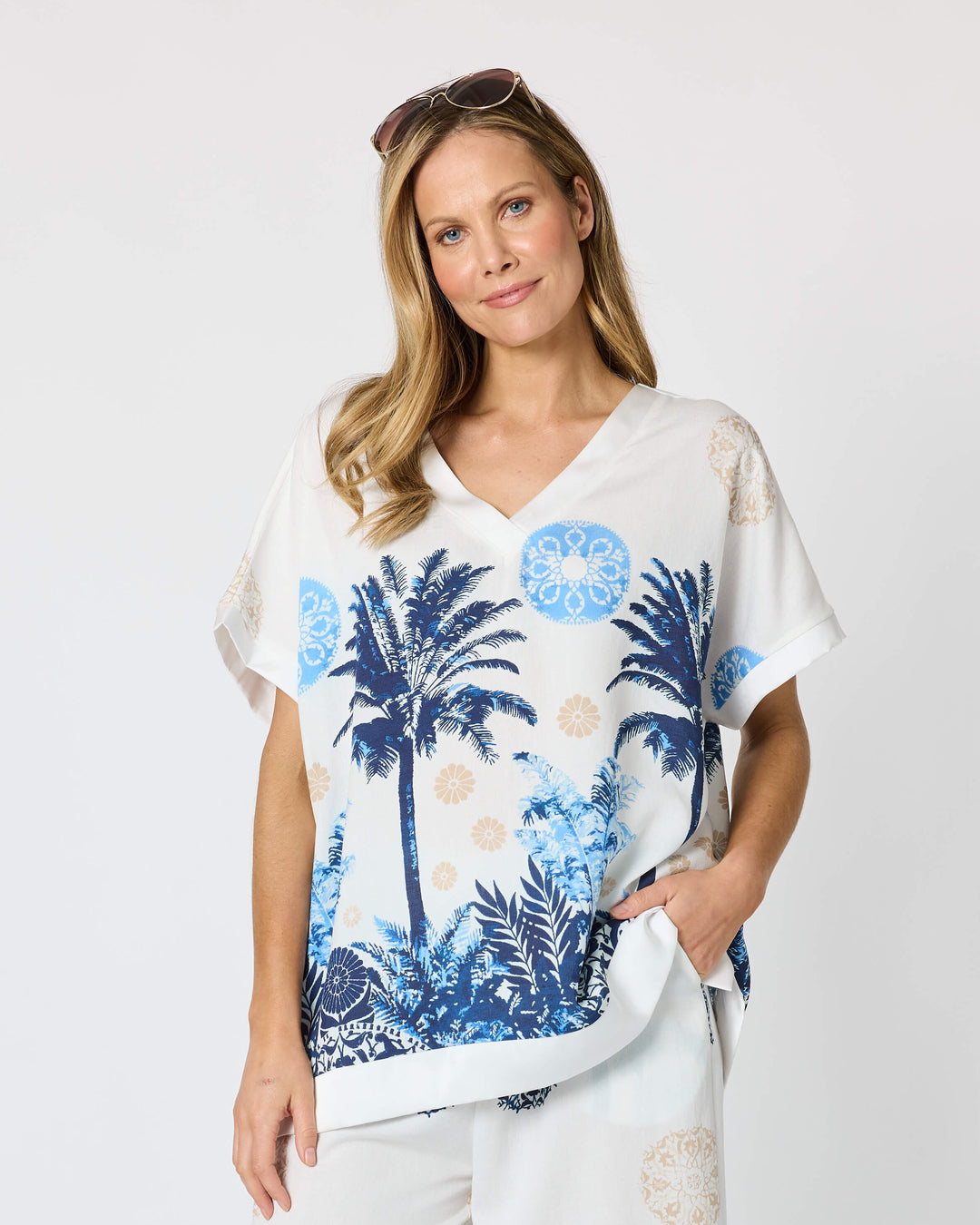 Clarity  woman modeling a matching two-piece lounge set featuring a white short-sleeve top and cropped pants with a blue tropical palm tree print, styled with beige slide sandals against a plain white studio background.