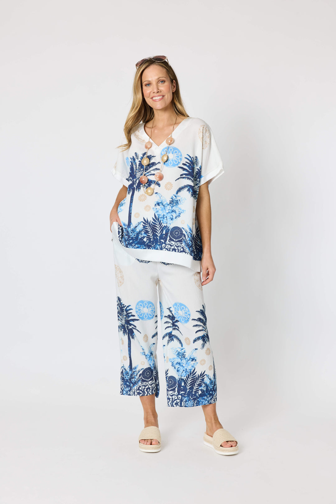 Clarity  woman modeling a matching two-piece lounge set featuring a white short-sleeve top and cropped pants with a blue tropical palm tree print, styled with beige slide sandals against a plain white studio background.