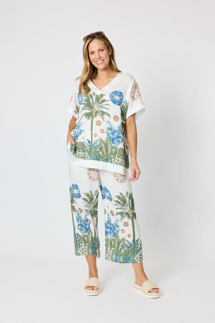 Clarity modeling a matching two-piece lounge set featuring a white base with a tropical print of palm trees, blue flowers, and green foliage, paired with white slide sandals against a light gray studio background.