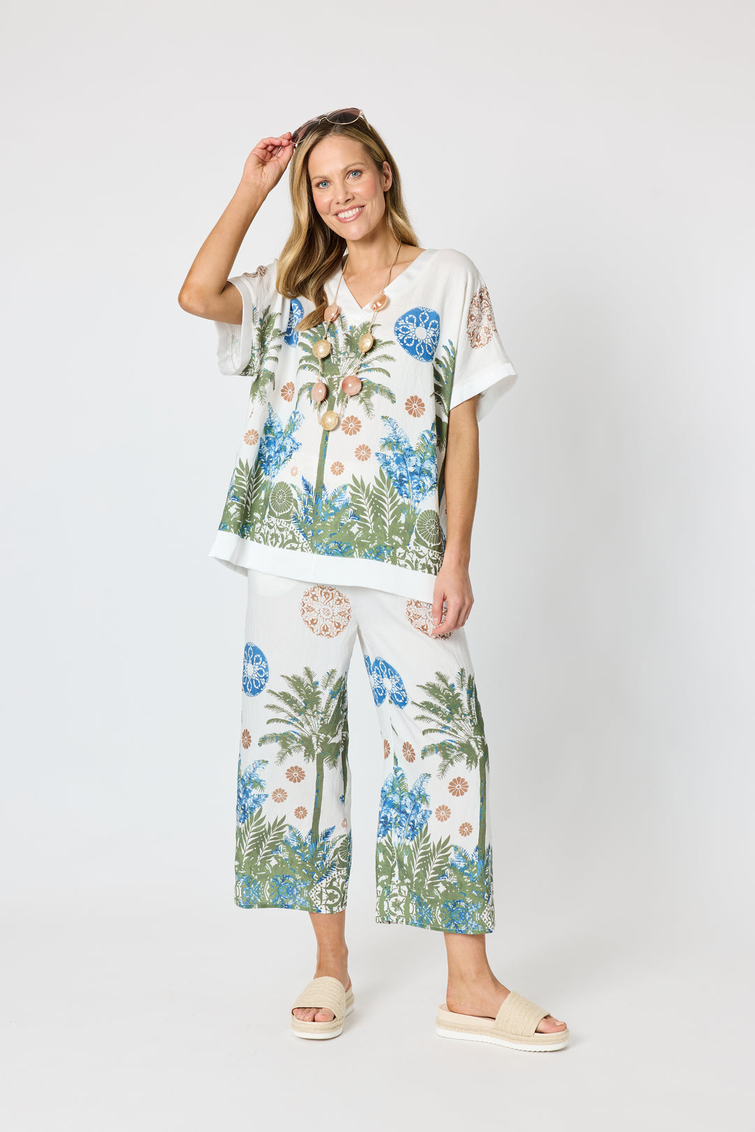 Clarity modeling a matching two-piece lounge set featuring a white base with a tropical print of palm trees, blue flowers, and green foliage, paired with white slide sandals against a light gray studio background.