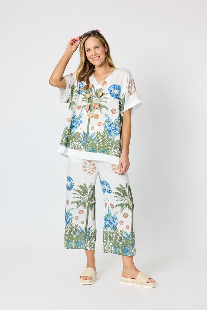 Clarity modeling a matching two-piece lounge set featuring a white short-sleeve top and wide-leg pants with a tropical palm tree and floral print in blue, green, and beige, set against a plain light gray studio background.