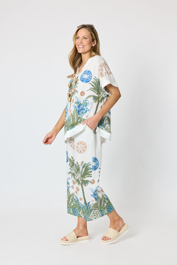Clarity modeling a matching two-piece lounge set featuring a white base with a tropical print of palm trees, blue flowers, and green foliage, paired with white slide sandals against a light gray studio background.
